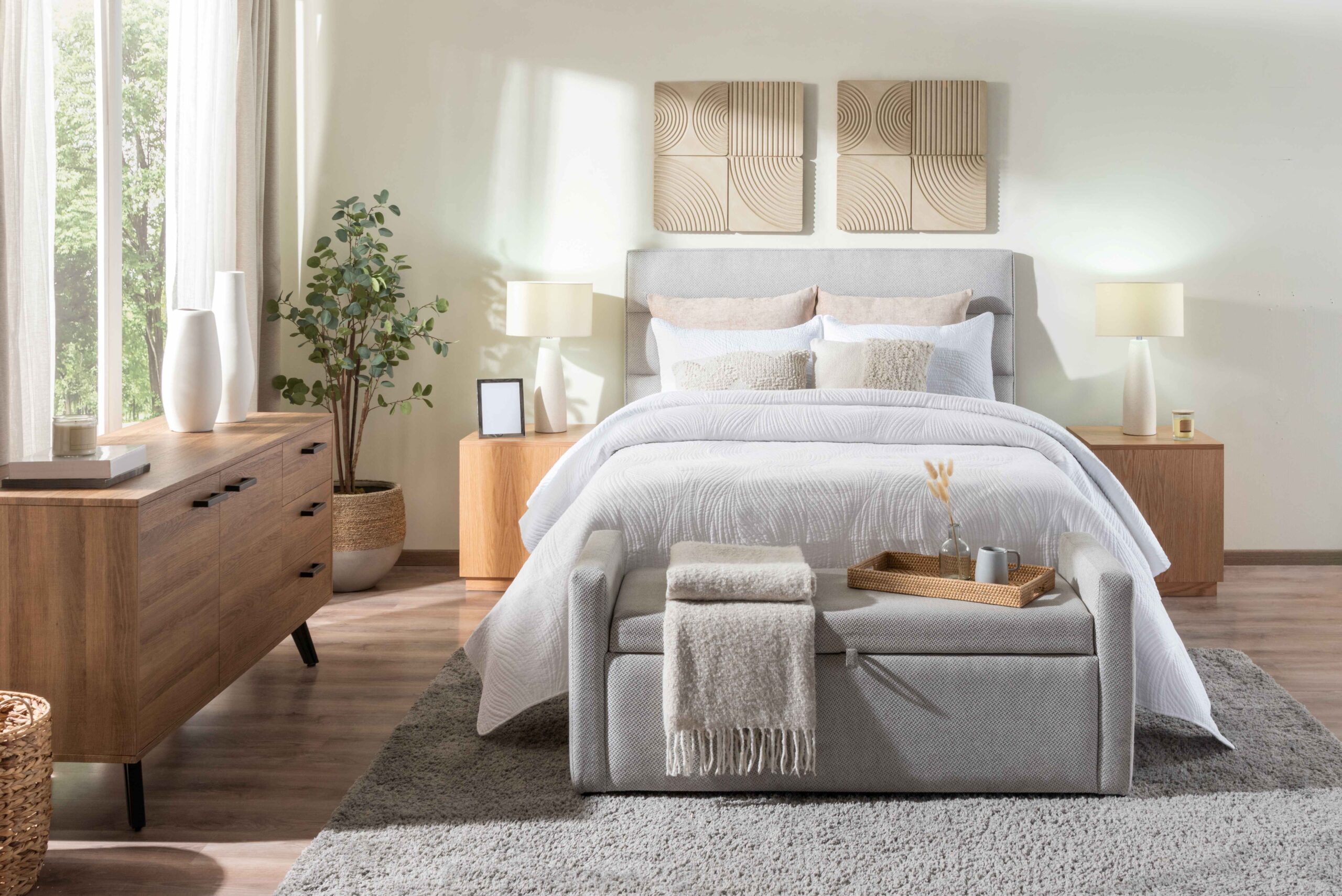 Japandi Schlafzimmer mit grauem gepolstertem Bett, weißer gesteppter Bettwäsche, Eichen-Nachtkästen und Kommode, Ottomane mit Tablett, gewebten Akzenten